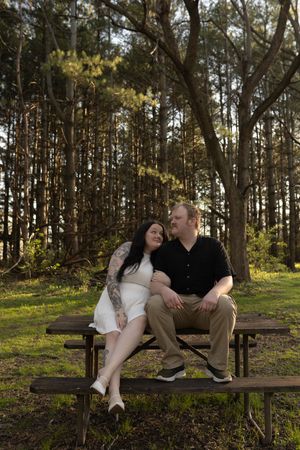 Couple sitting close on a picnic table in a forest, smiling at each other, woman in white dress with tattoos.