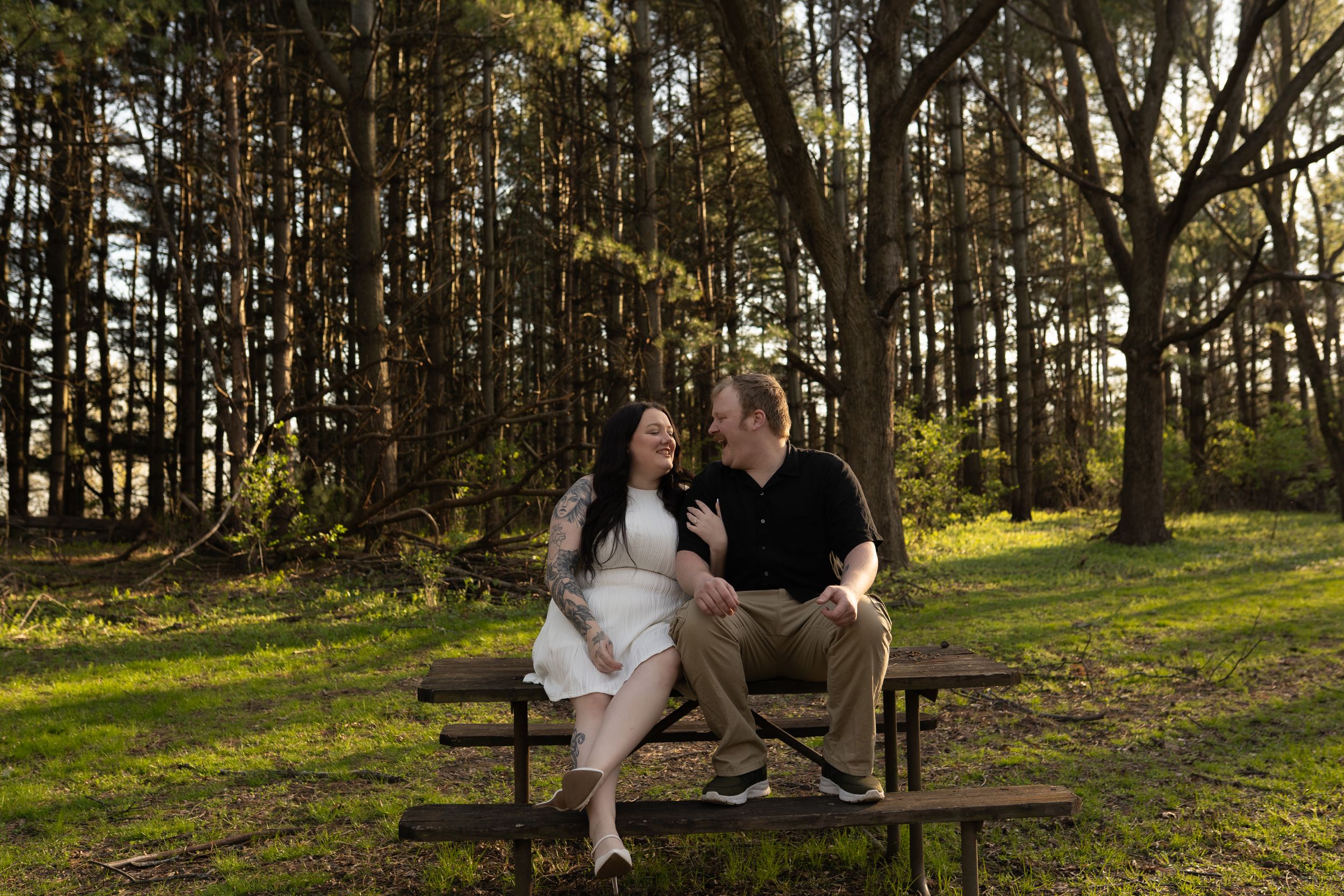 Couple sitting together on a picnic table in a lush green forest park, smiling at each other during an engagement photo.