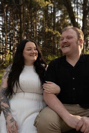 A couple smiles together in a forest, woman in white dress with tattoos and man in black shirt holding hands.