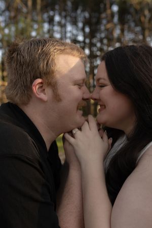 A couple shares a tender romantic moment, nearly kissing while holding hands outdoors among trees.