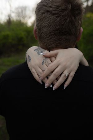 Tattooed woman with engagement ring embraces man in black shirt from behind in outdoor setting.