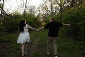 A couple walks hand-in-hand down a wooded path, woman in white dress and man in black shirt at dusk.