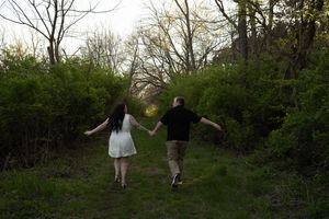 Couple holding hands walking through lush green forest path, woman in white dress and man in black shirt.