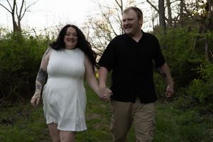 Couple holding hands and smiling while walking outdoors, woman in white dress with tattoos, man in black shirt.