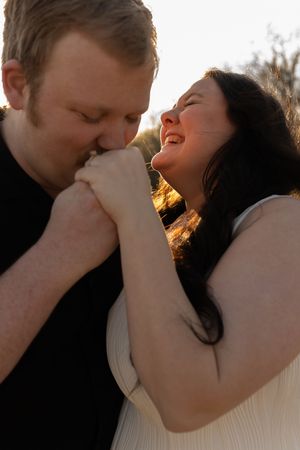 A man kisses a laughing woman's hand tenderly in warm golden sunset light outdoors.
