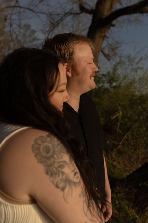 Couple in golden hour light outdoors, woman with sunflower tattoo leaning against man in black shirt.