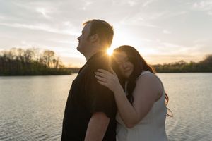 Romantic couple standing by a lake at golden sunset, woman leaning on mans shoulder smiling.