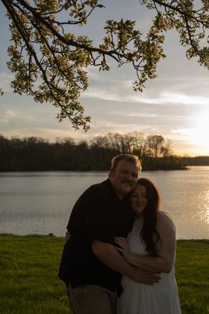 Engaged couple embracing under flowering tree branch by a lake at sunset on green grass.