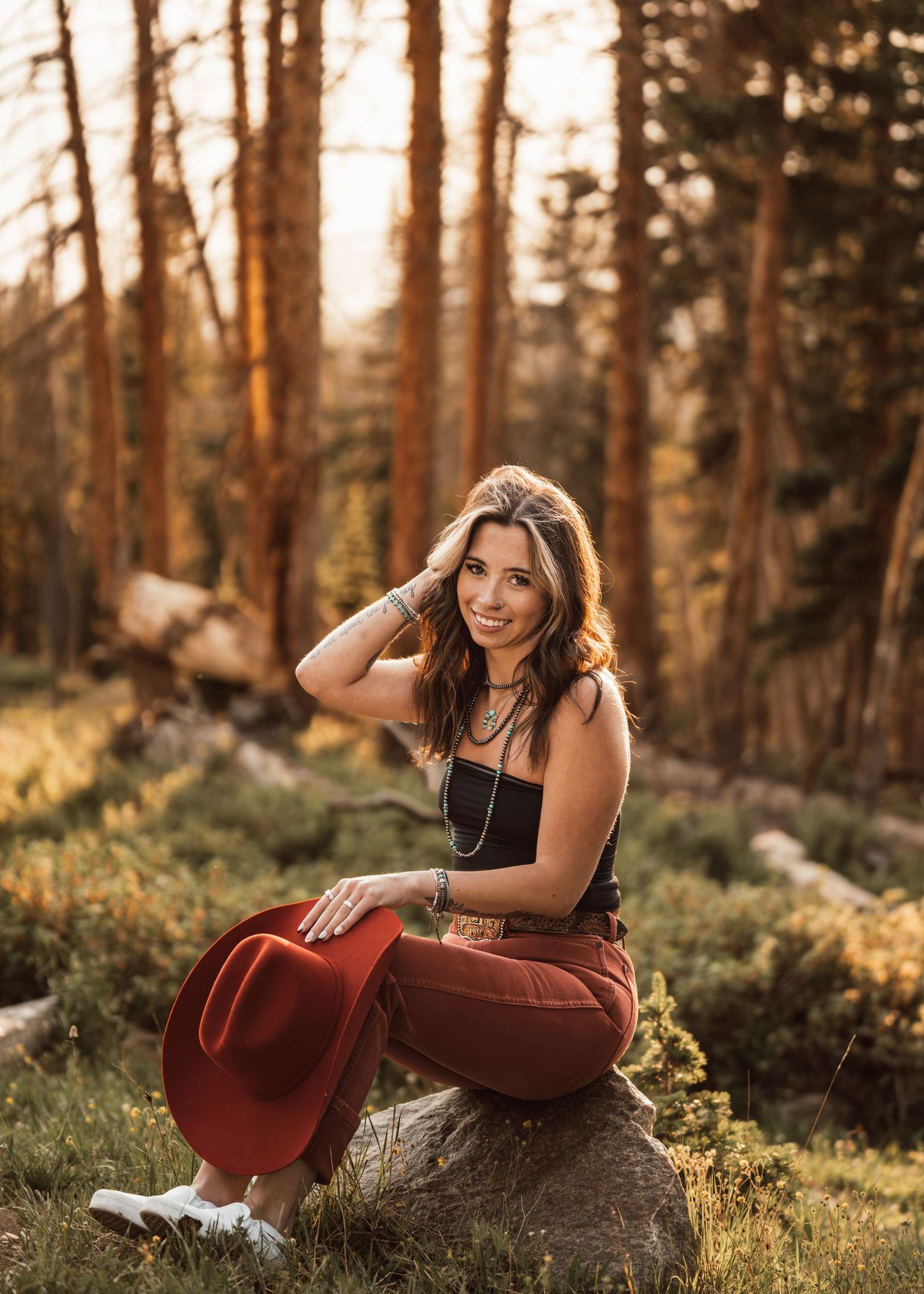 Young woman in black cowboy hat, striped shirt, crop top, and flare jeans smiling near a mountain lake at sunset.