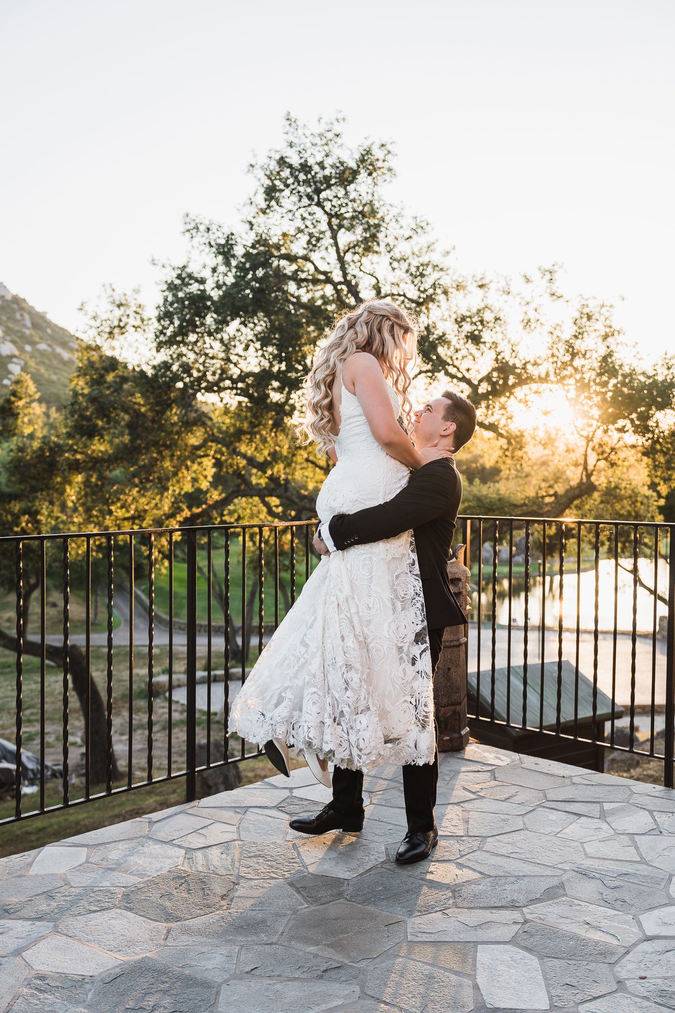 Groom lifting bride in lace wedding dress at sunset on stone terrace with trees and mountains in background.