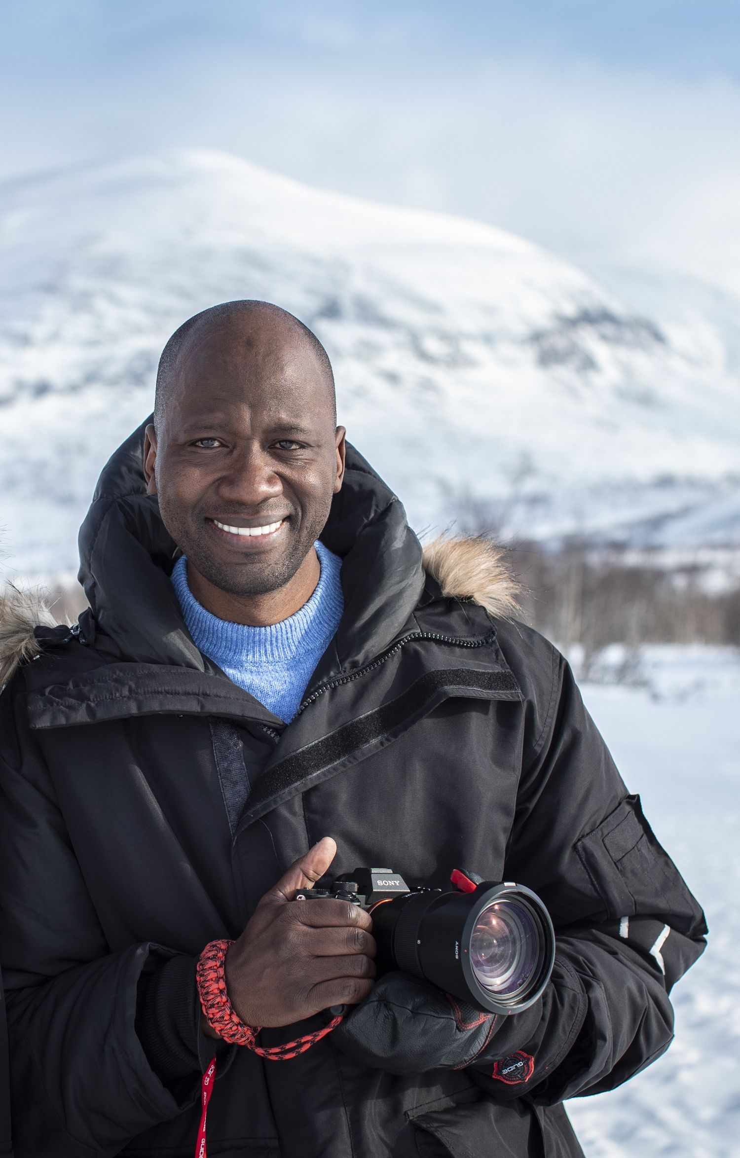 Smiling man holding a camera in a snowy mountain landscape wearing a black winter jacket.