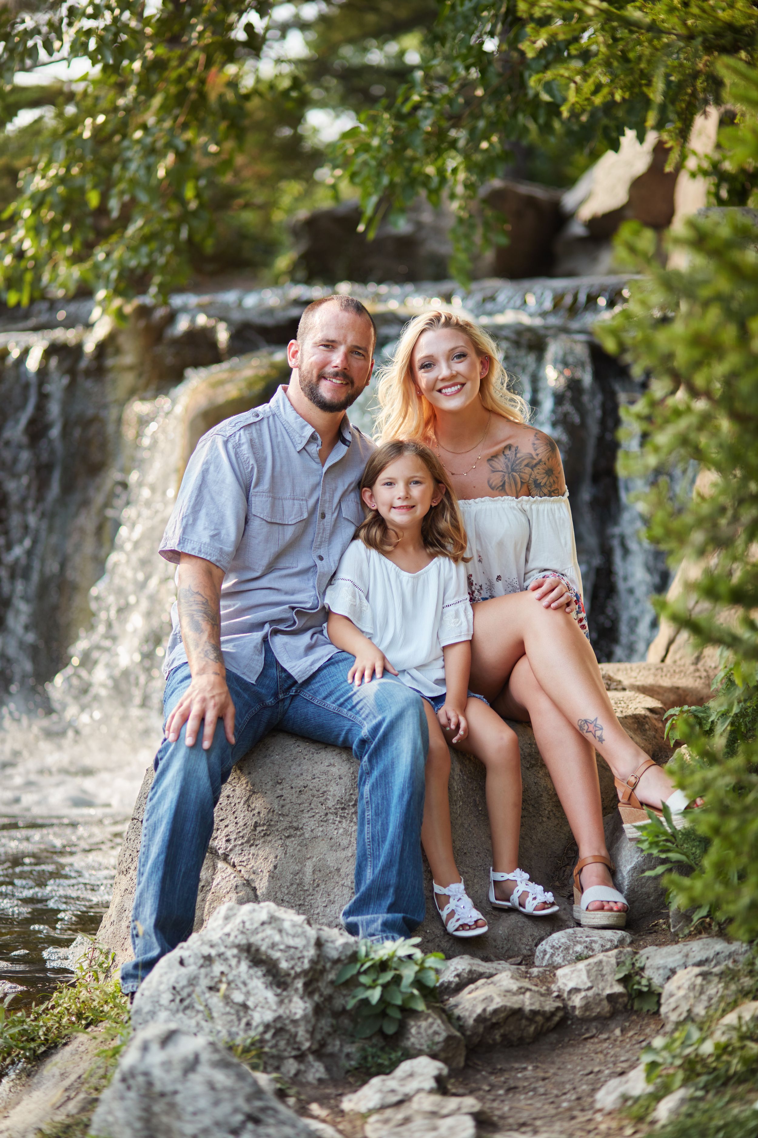 Happy family of three sitting on rocks near a waterfall, smiling outdoors surrounded by lush green trees.