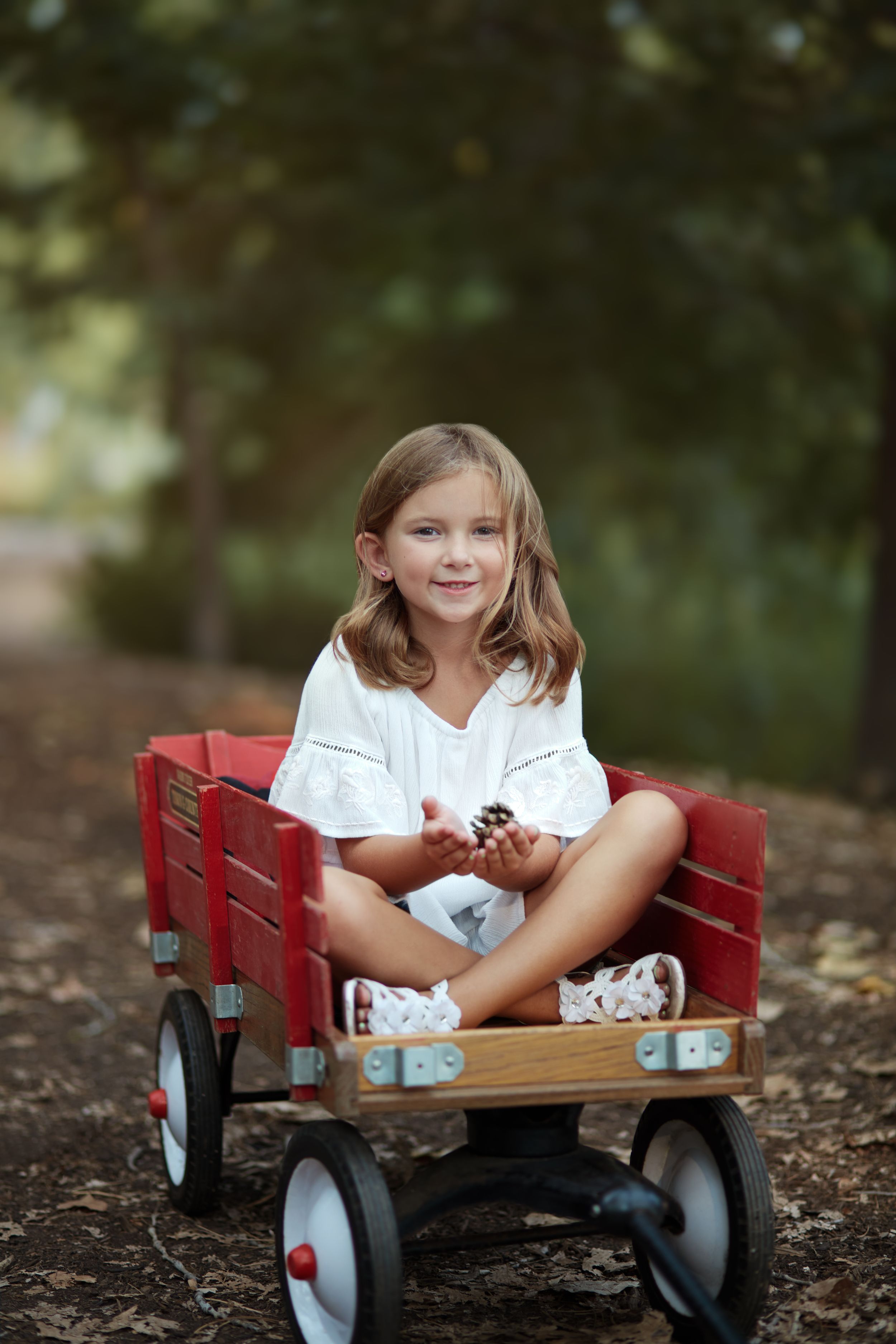 Young girl sitting cross-legged in a red wooden wagon outdoors, smiling and holding a pinecone.