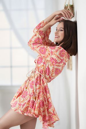 Young woman in pink floral mini dress posing gracefully against white wall, smiling with hand raised.