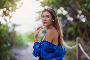 Young woman in blue off-shoulder top poses on a scenic outdoor path surrounded by lush green trees.