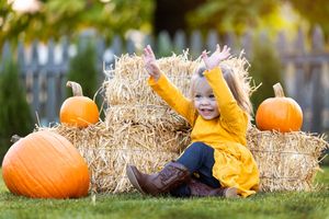 Smiling toddler girl in yellow dress sitting on hay bale with orange pumpkins in fall outdoor setting.