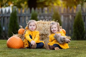Two young girls in yellow outfits sit by hay bales and pumpkins in an autumn backyard setting.