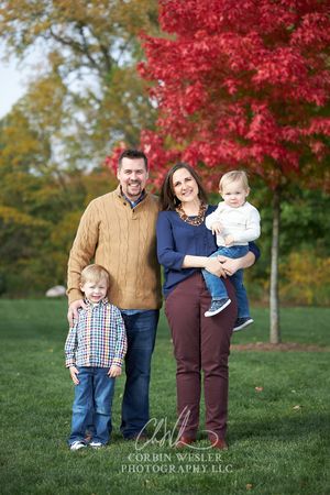 Family of four posing outdoors in fall with vibrant red maple tree, parents with two young boys smiling in autumn park.