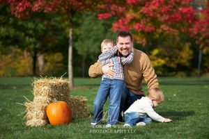 Father laughing with two young children outdoors in fall, hay bales and pumpkin nearby, colorful autumn trees in background.