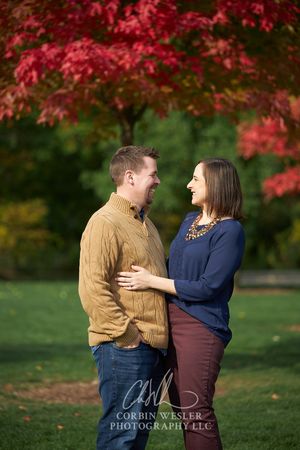 Couple laughing together outdoors in autumn park with vibrant red maple tree foliage backdrop during fall engagement photo session.