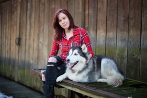 Young woman in red plaid shirt sits with Alaskan Malamute dog on wooden bench near rustic fence.