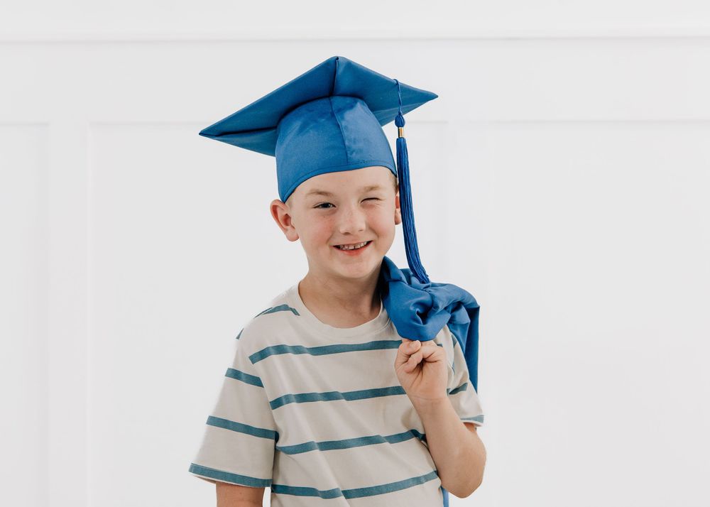 Smiling young boy wearing a blue graduation cap and gown, posing proudly against a white background.