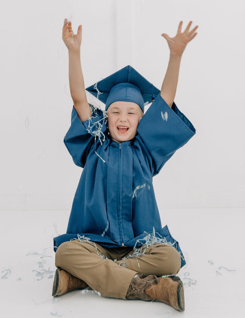 Joyful child in blue graduation cap and gown sitting cross-legged with arms raised, celebrating with confetti.