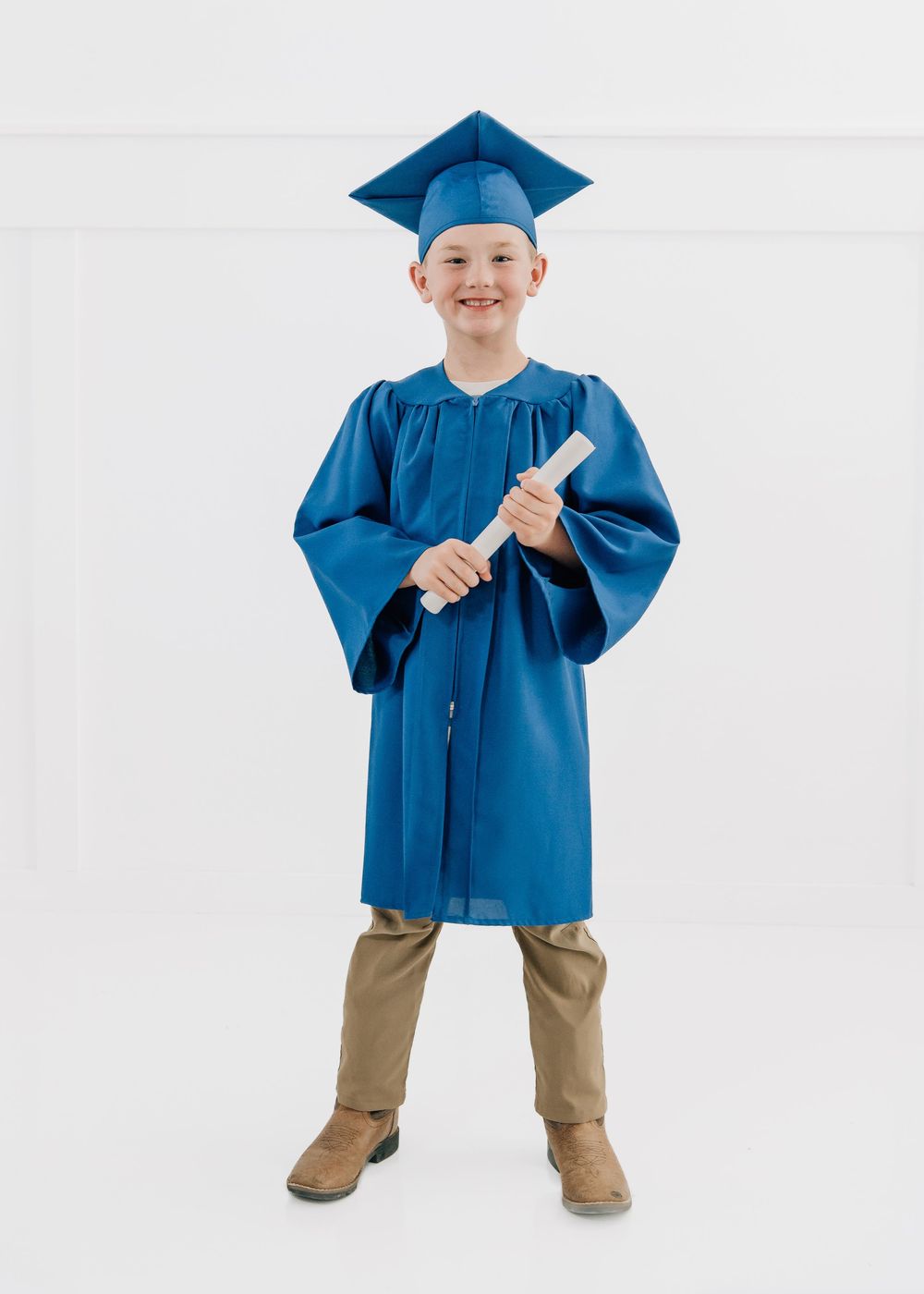 Young boy wearing a blue graduation cap and gown, smiling and holding a diploma scroll.