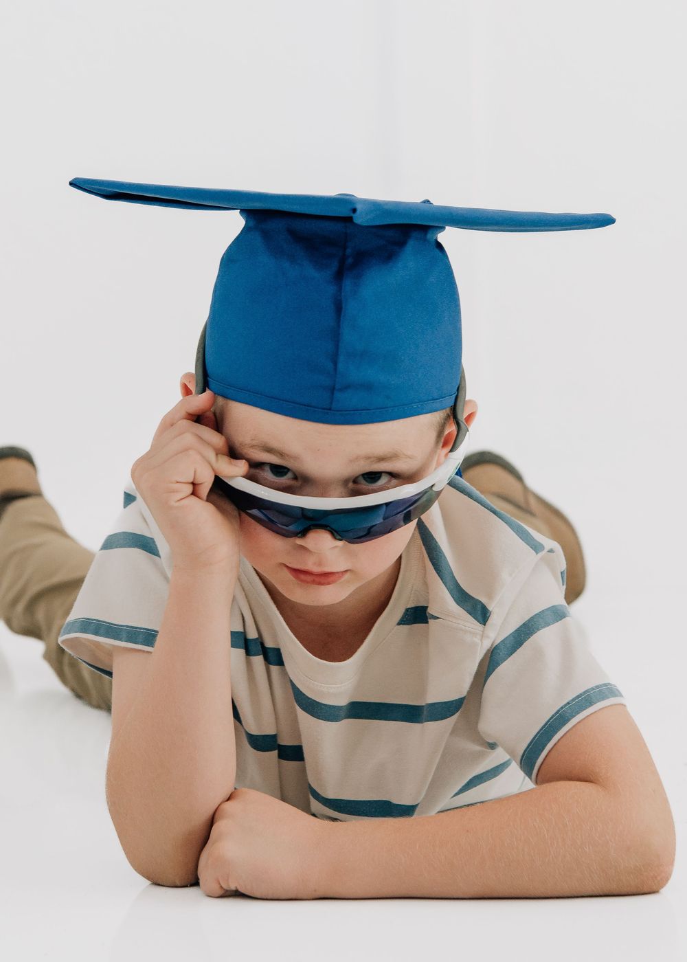 A young boy wearing a blue graduation cap and sunglasses lies on the floor with a serious expression.