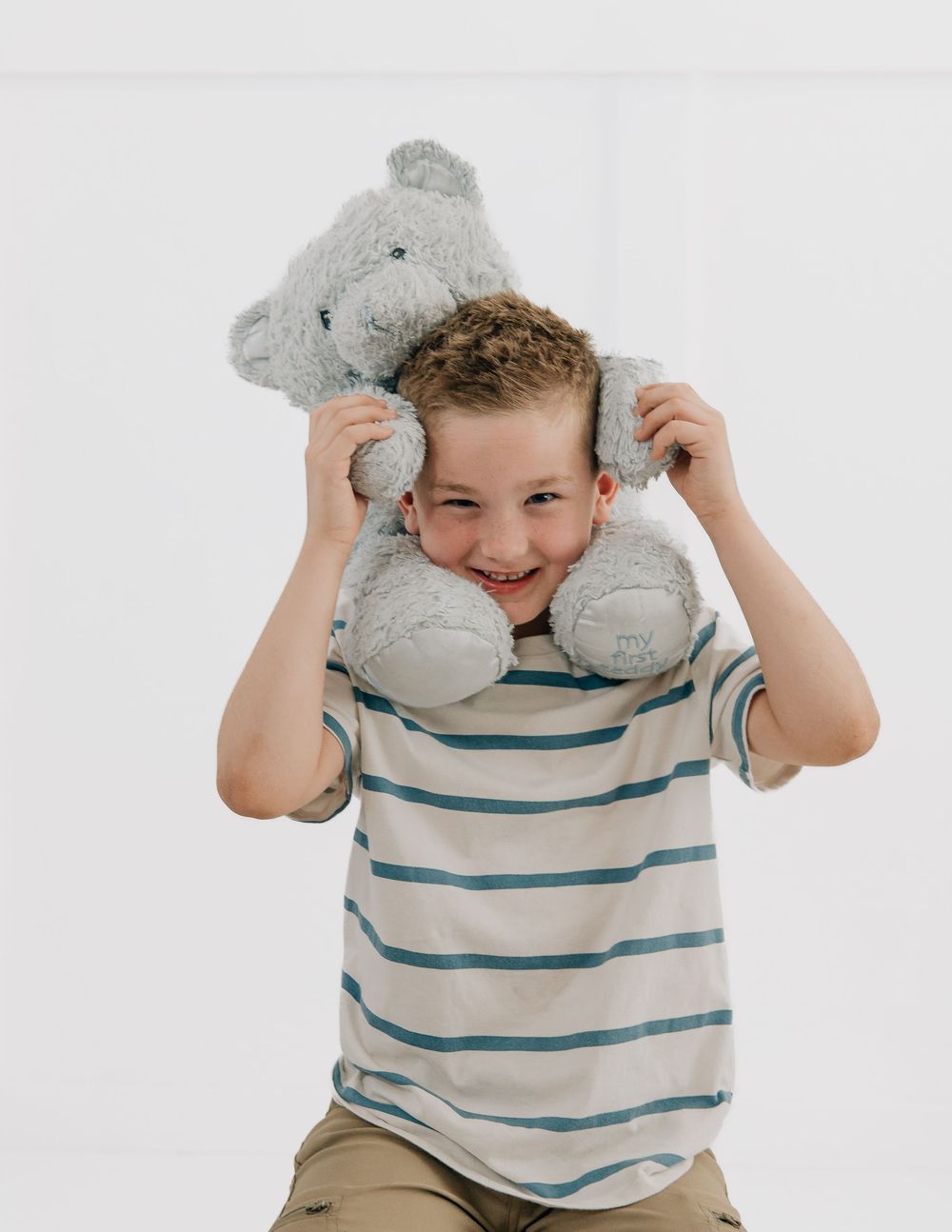 Smiling boy in striped shirt holding a gray teddy bear above his head against a white background.