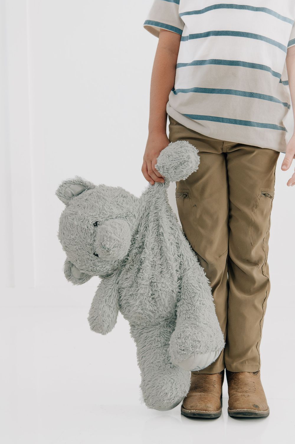 Child holding a gray teddy bear stuffed animal while wearing striped shirt, khaki pants, and brown boots.