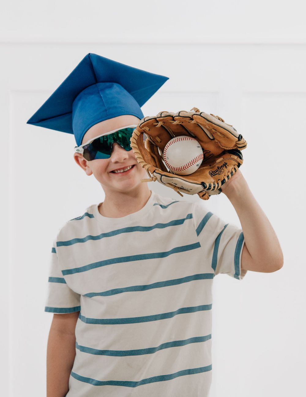 Boy in blue graduation cap and sunglasses holding baseball glove with ball, wearing striped t-shirt.