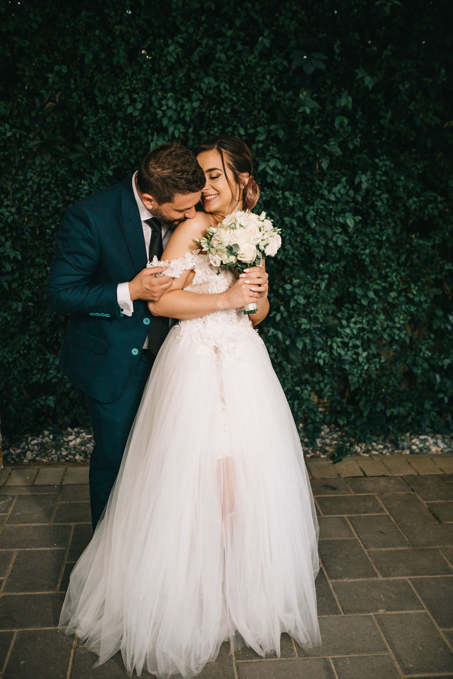 Bride in white tulle gown holding bouquet embraced by groom in navy suit against lush green ivy wall.