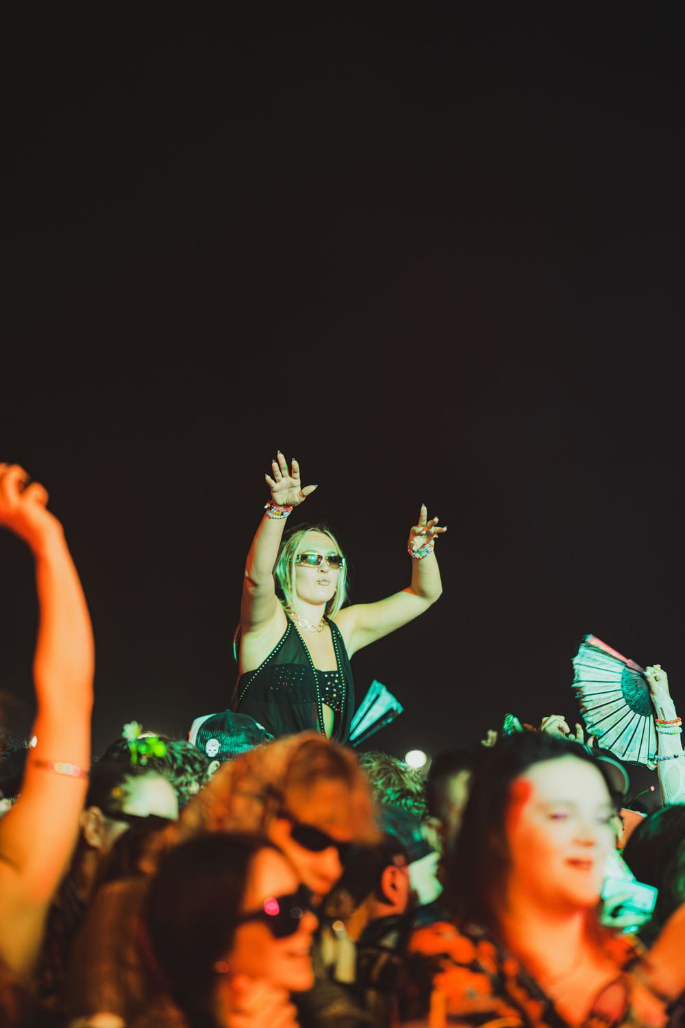 Excited woman raises hands in crowd at outdoor night music festival, wearing sunglasses under colorful stage lights.