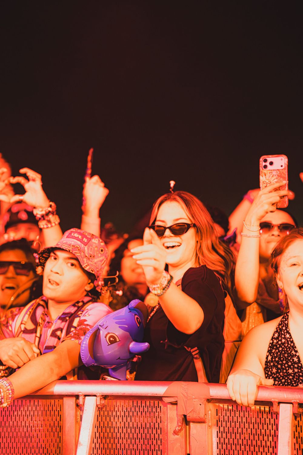 Excited concert crowd at night, woman in sunglasses clapping and smiling, holding a stuffed animal at a music festival.