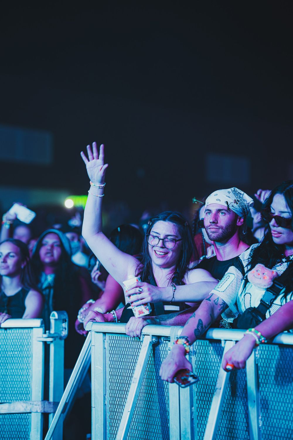 Excited concert crowd waves at music festival barrier under blue stage lights at night.