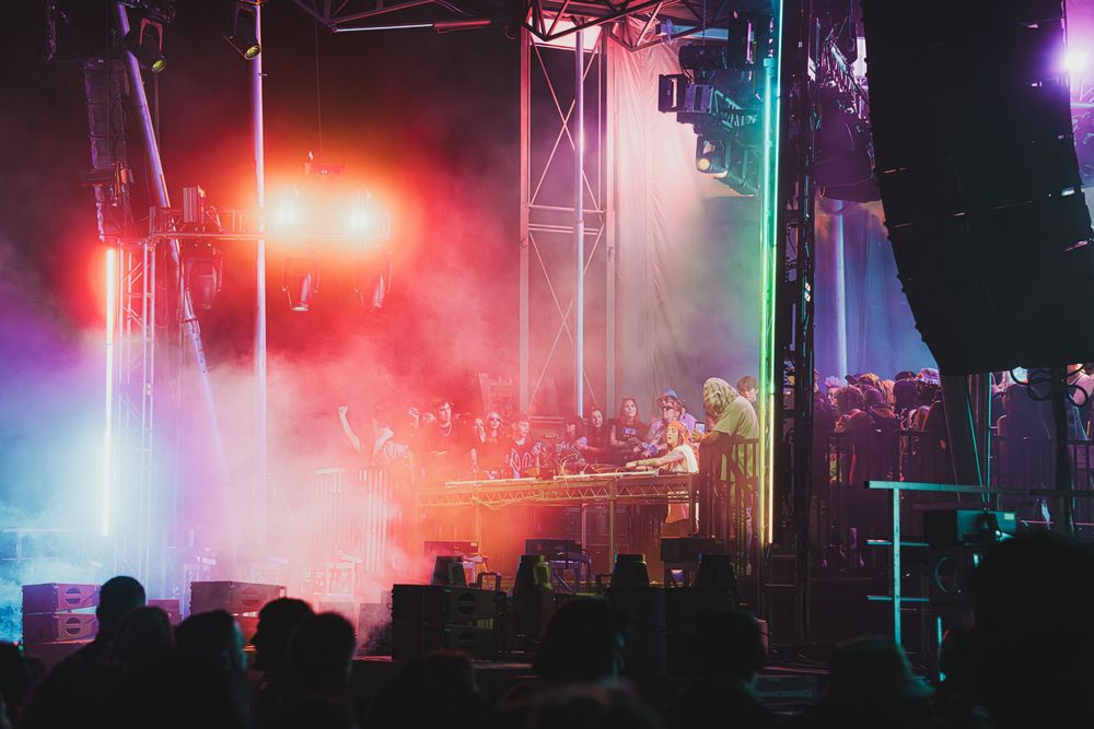 Live concert stage with colorful red and green lights, fog effects, and silhouetted crowd at an outdoor music festival.