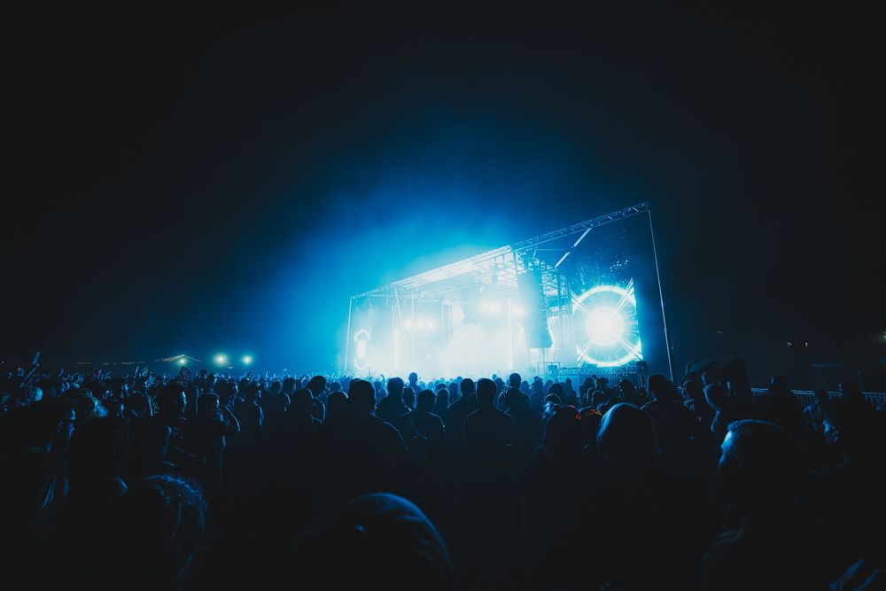 Large outdoor music festival crowd at night with bright blue stage lights illuminating the dark sky.