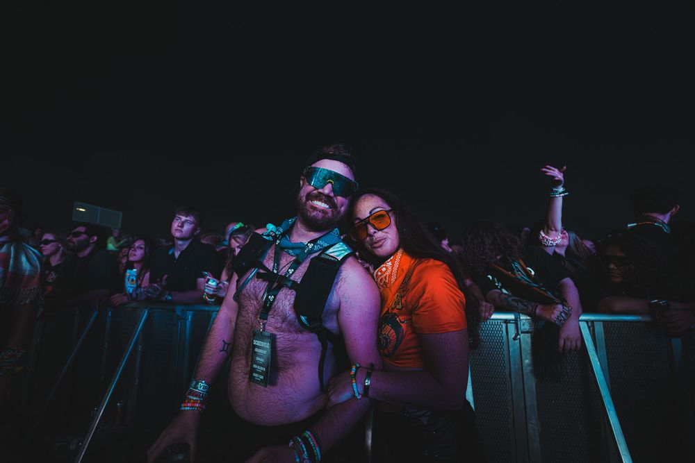 Two festival-goers pose and smile at an outdoor music festival at night under colorful neon lights.