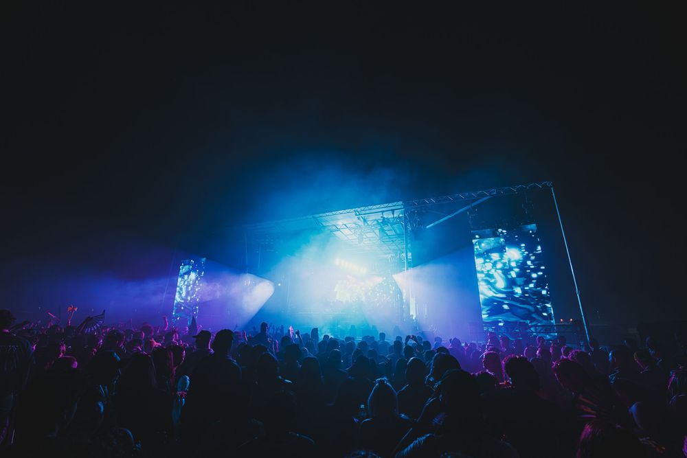 Massive outdoor music festival stage illuminated by bright blue and purple lights with large crowd.