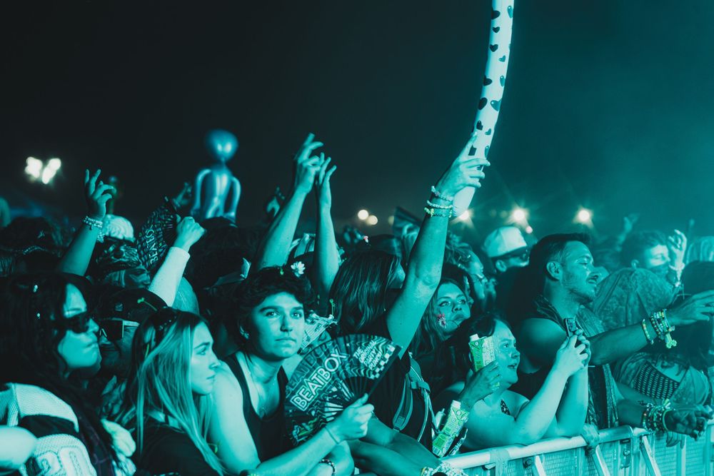 Excited concert crowd under green lights, fans cheering and raising hands at an outdoor music festival at night.