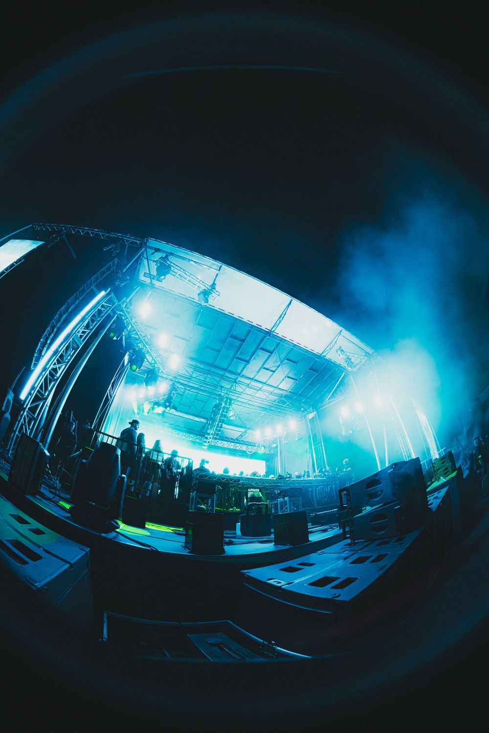 Fisheye lens view of a concert stage with blue lighting, fog, and scaffolding at an outdoor music festival.