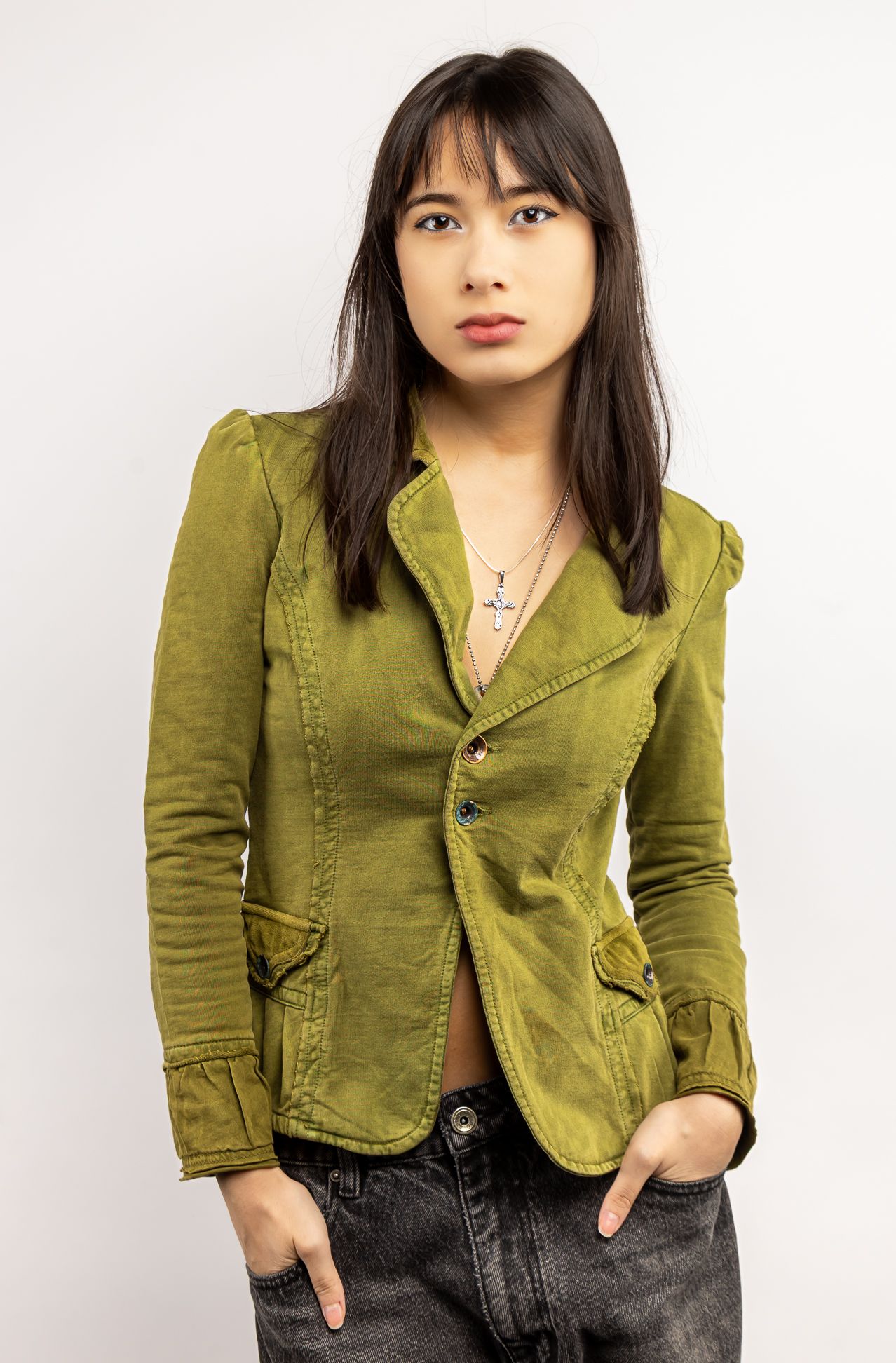 Girl in front of a white backdrop in a green button-up blouse, and black jeans with her hands in her pockets, looking straight at the camera