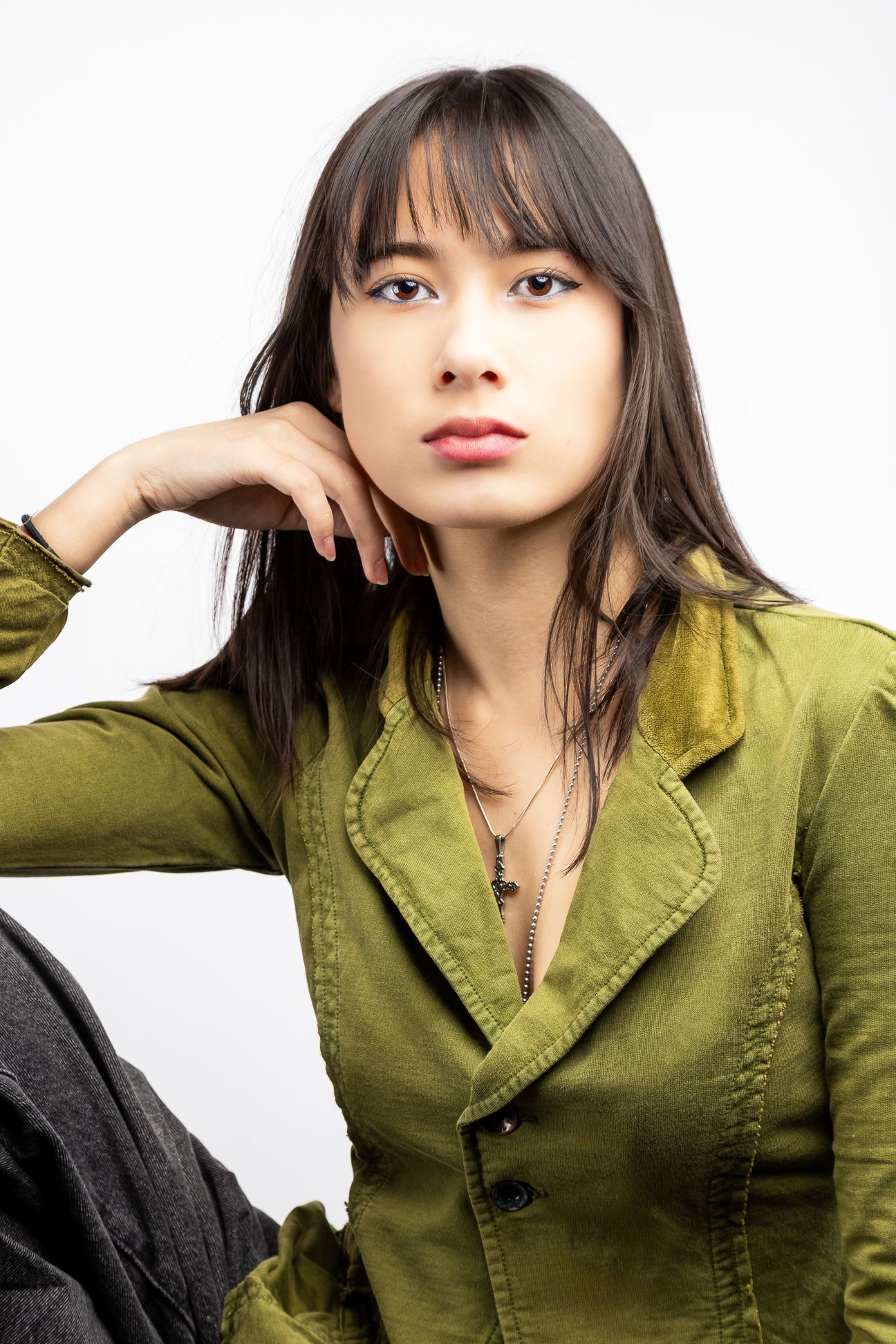 Girl in front of a white backdrop in a green button up blouse, resting her head on her hand and looking straight