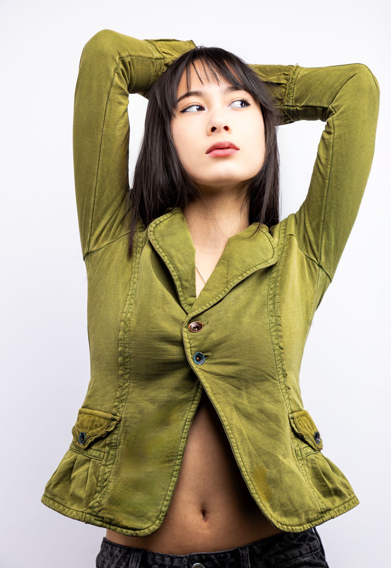 Girl in front of a white backdrop in a green button-up blouse, hands above her head and looking right