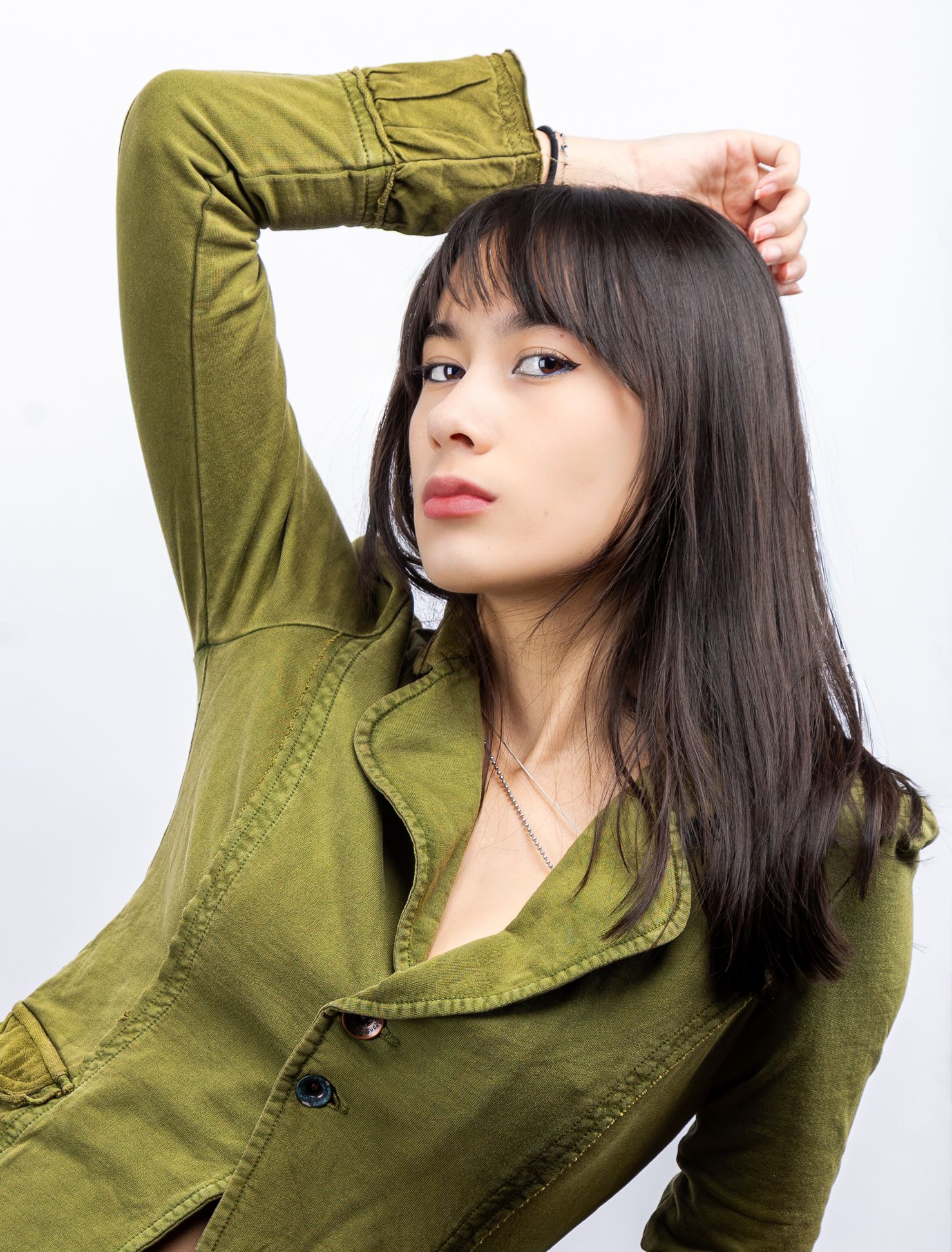 A girl in a green button-up top, looking at the camera from the side with one of her arms resting on her head