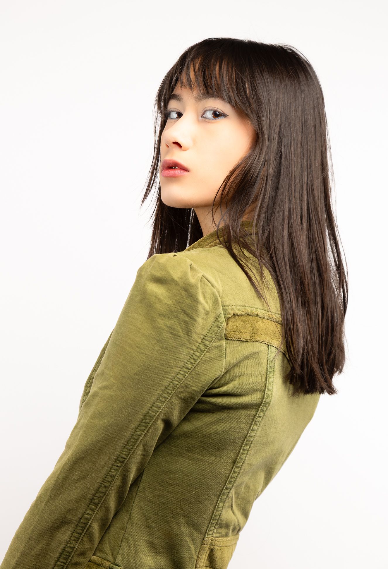 Girl in front of a white backdrop in a green button-up blouse, looking behind to look at the camera