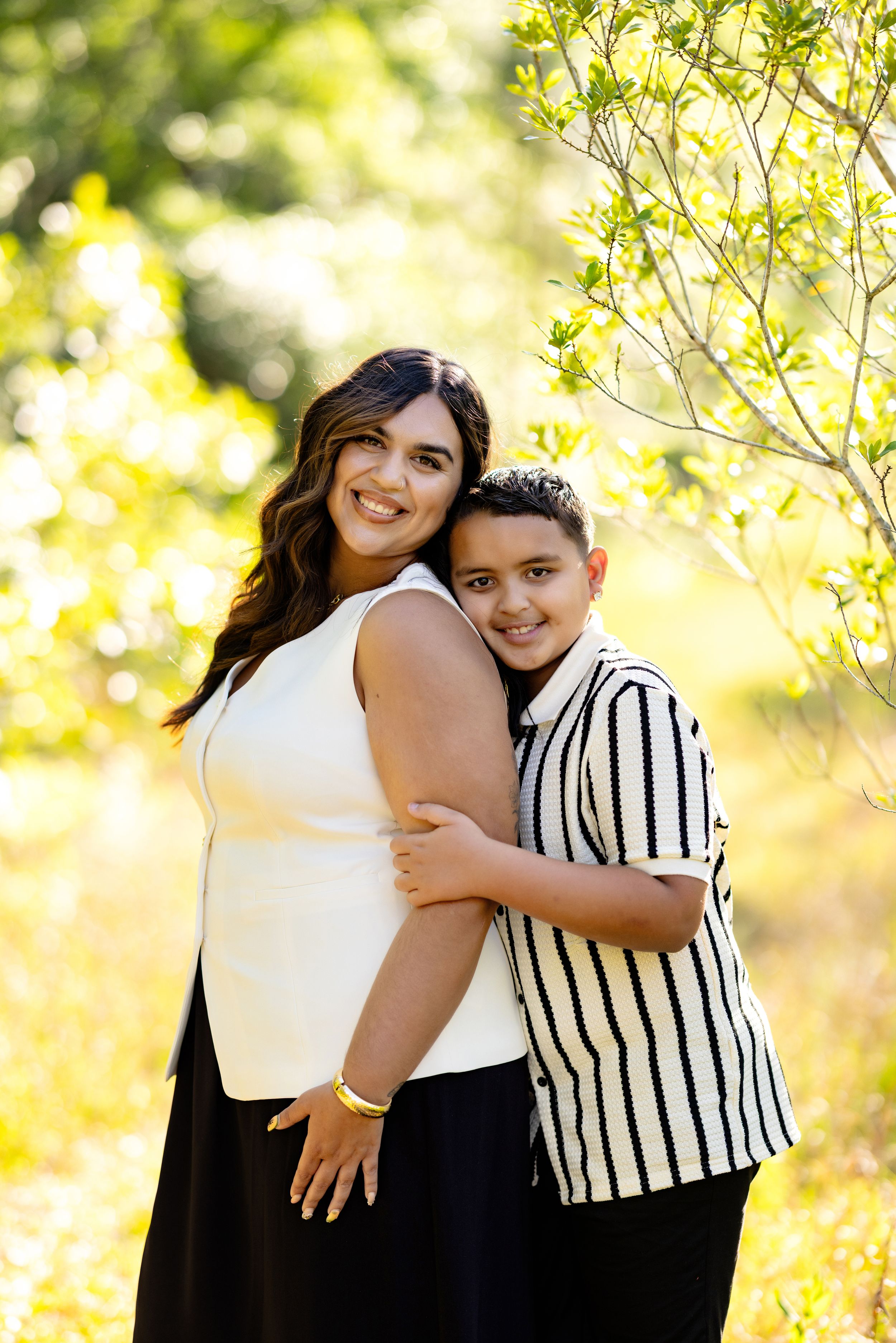A woman and young boy share a warm embrace outdoors, smiling together among lush green trees in natural sunlight.