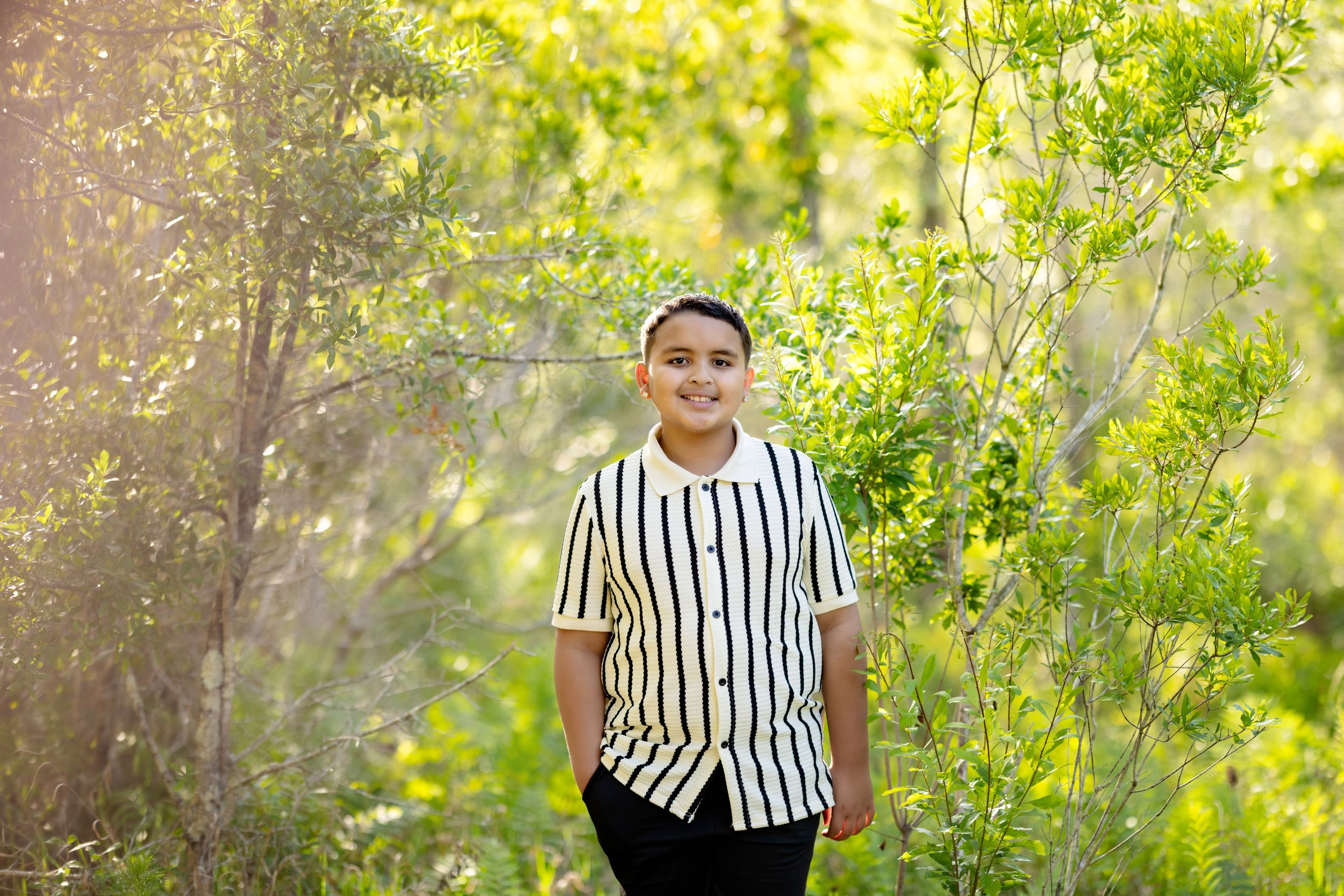 Smiling boy in striped shirt standing among lush green trees and shrubs in a sunlit outdoor setting.
