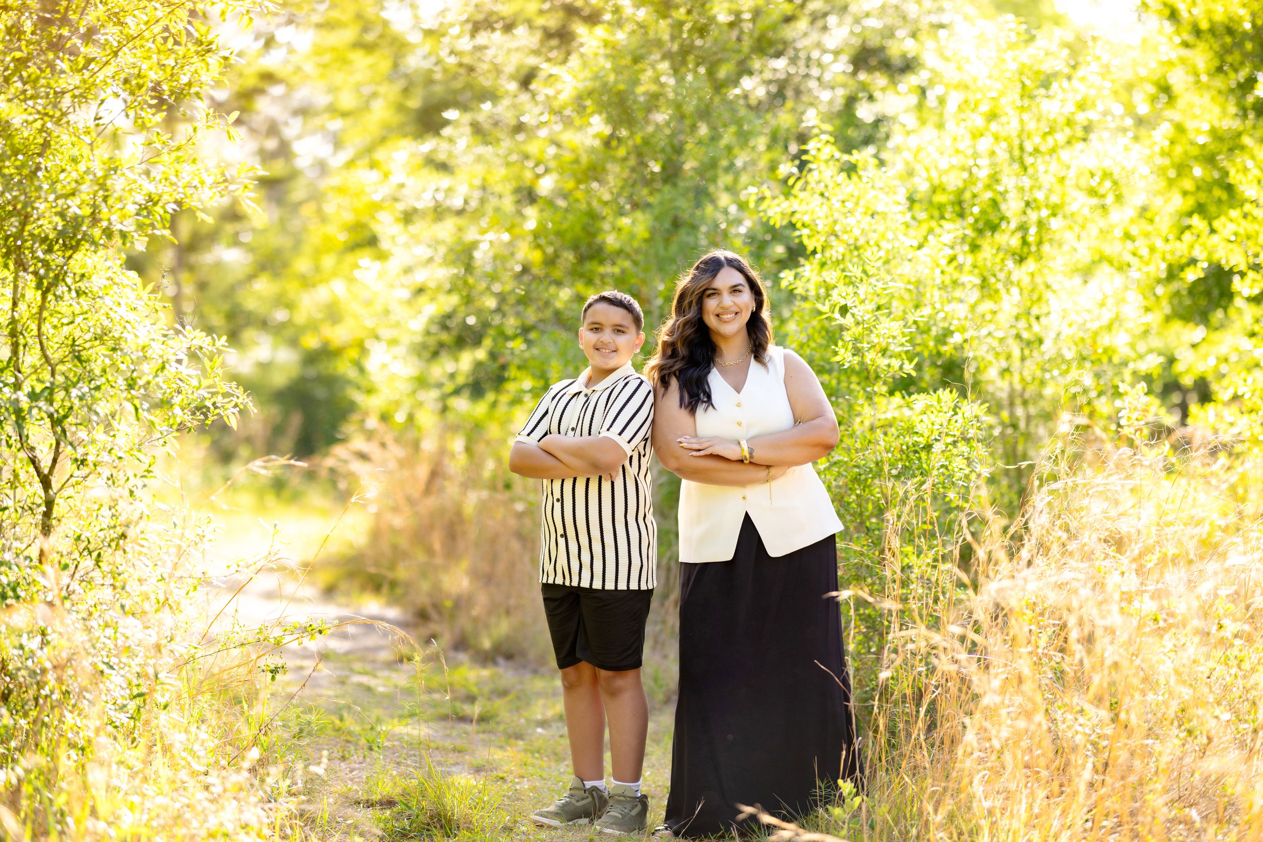 Two people standing on a sunlit nature path surrounded by lush green trees and golden grass on a bright day.